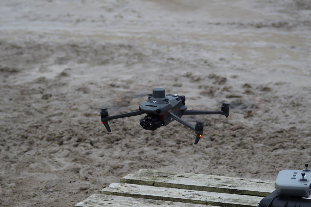 Eine graue Drohne fliegt über matschigem Boden mit einem Controller im Vordergrund.